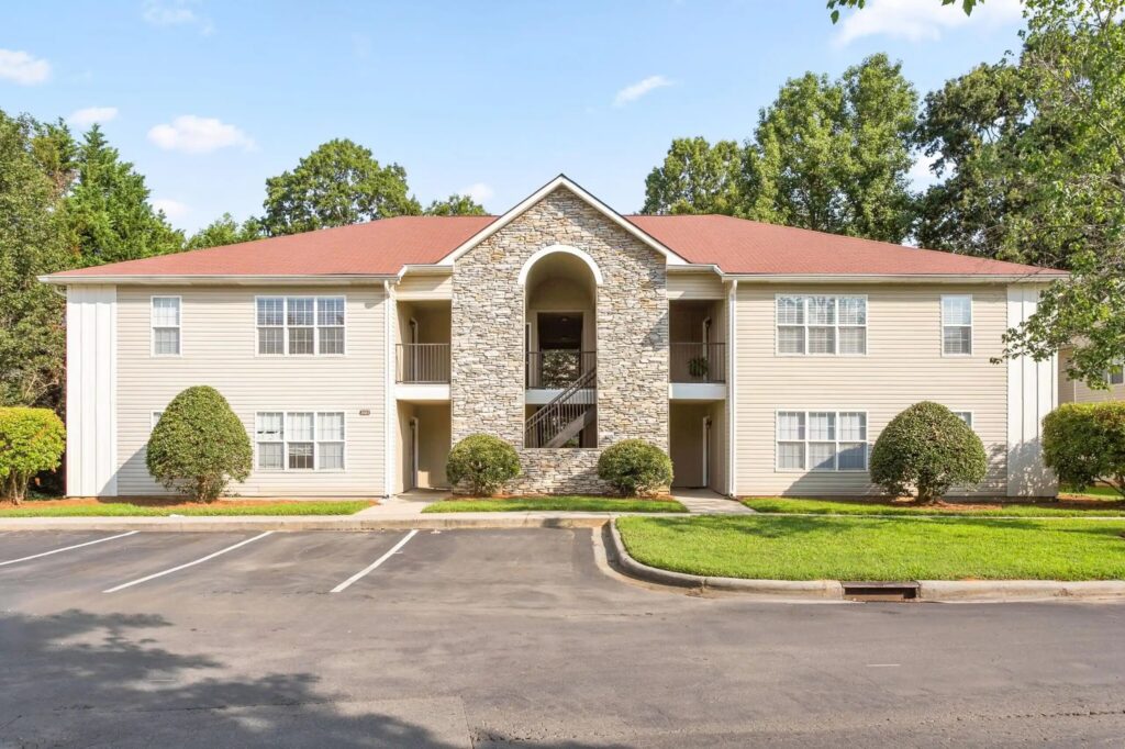 Two-story beige apartment building with red roof, central stone archway, surrounded by green grass and trees. Empty parking lot in the foreground.