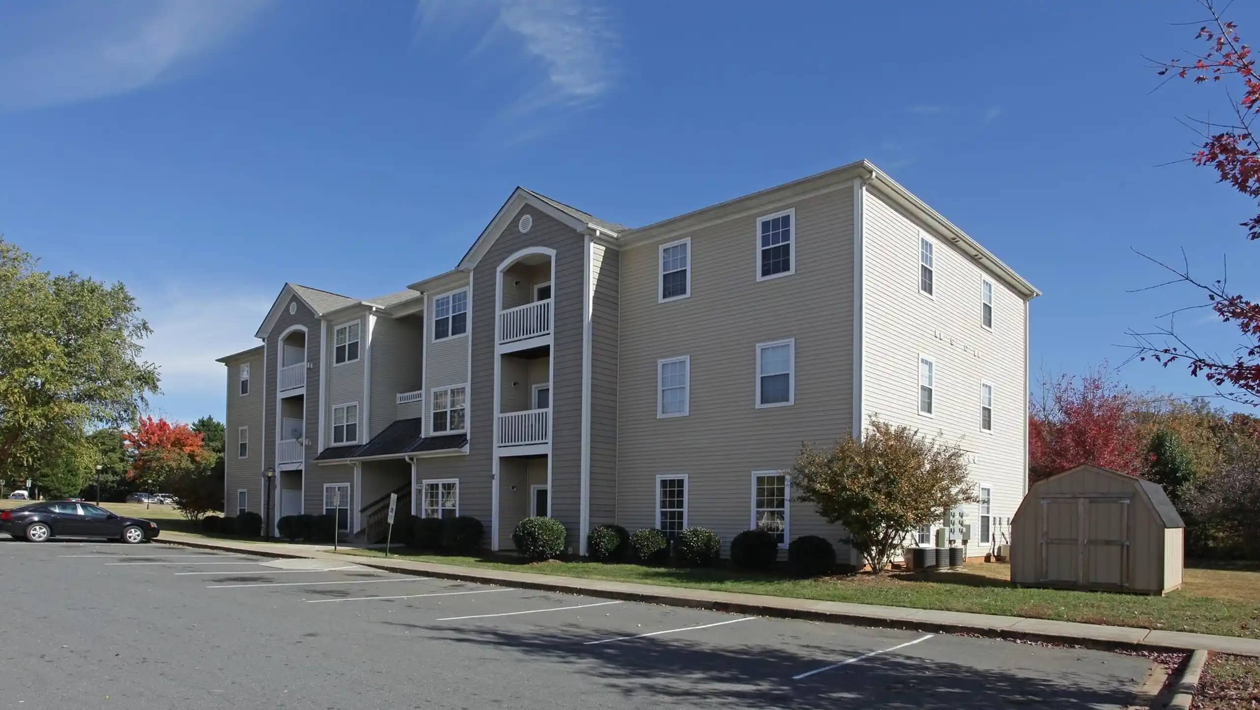 A beige, multi-story apartment building with balconies and a small shed nearby, set against a clear blue sky. An empty parking area is in the foreground.