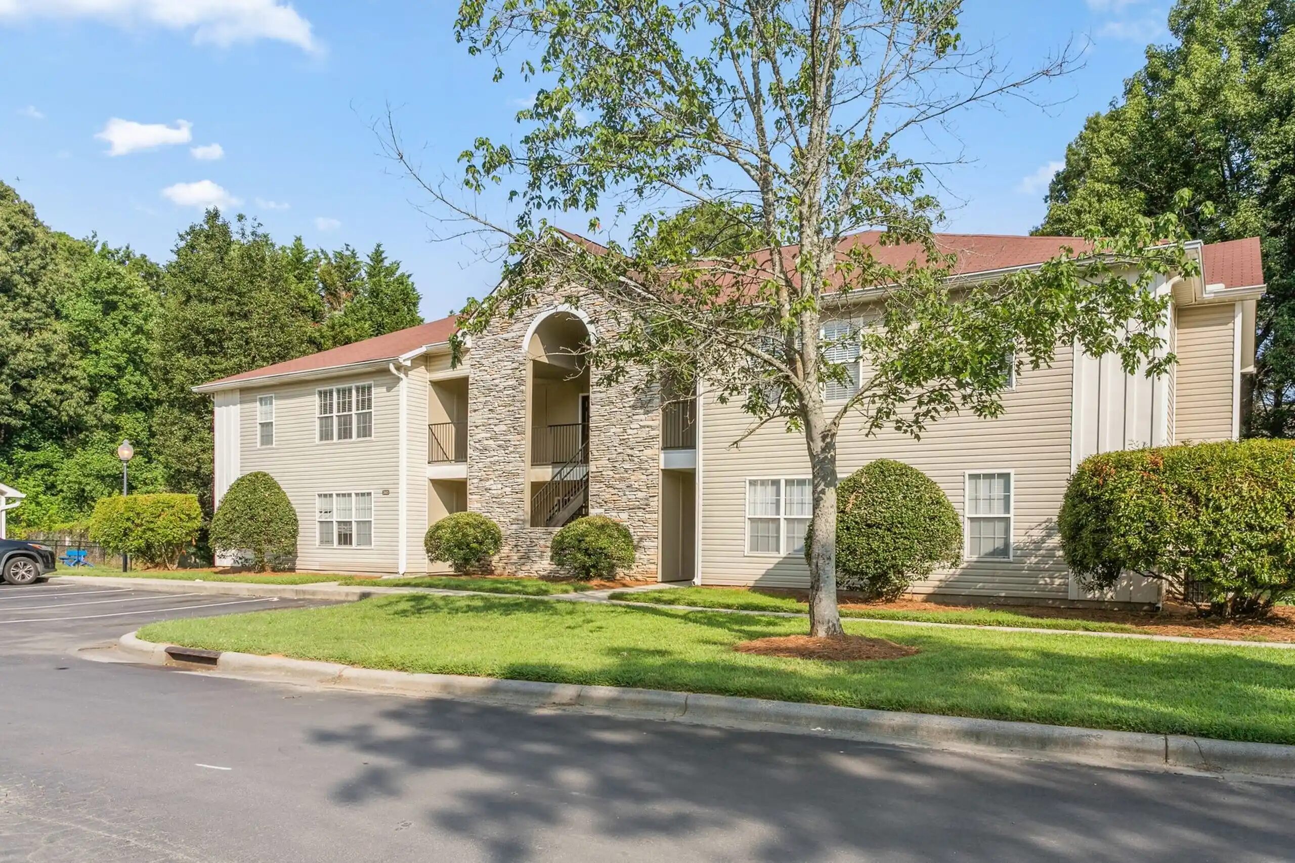 Two-story beige apartment building with a stone entrance, surrounded by green grass and trees. The sky is clear and blue.