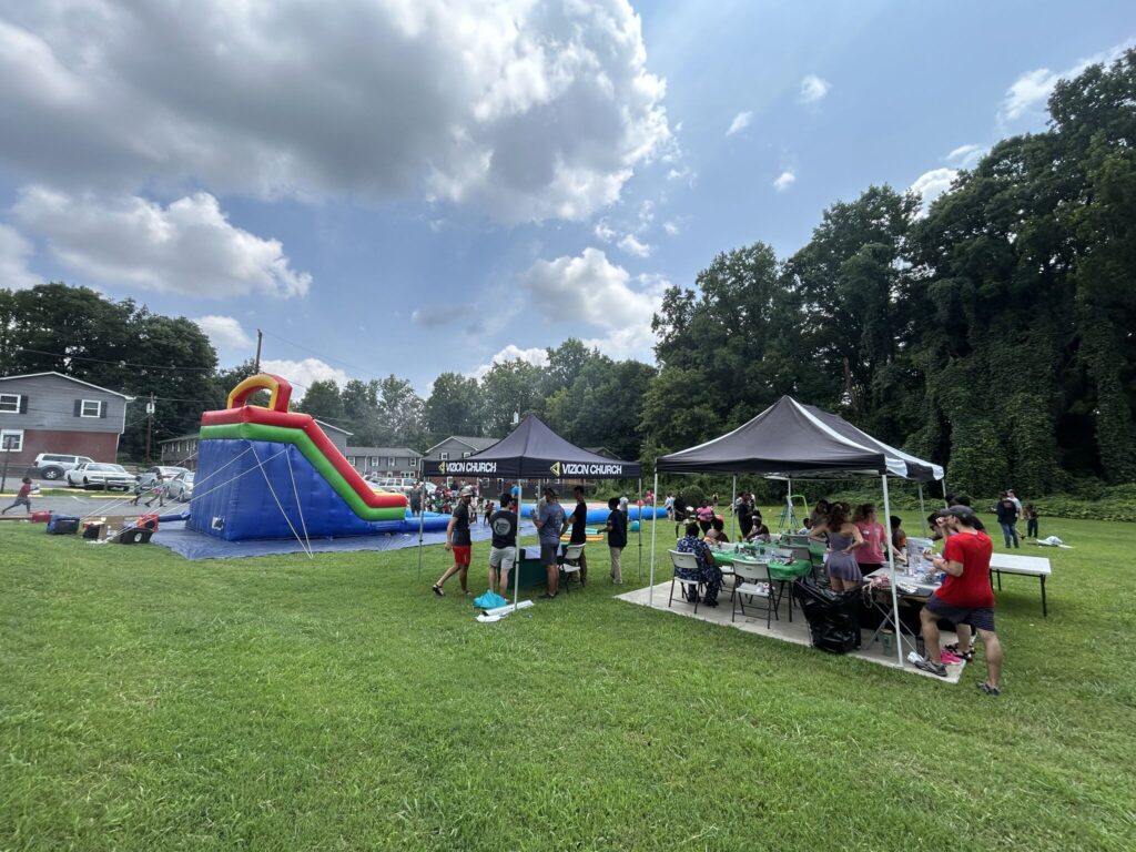 Outdoor gathering with a large inflatable water slide, people under tents, and picnic tables on a grassy area. Trees and houses are visible in the background under a partly cloudy sky.