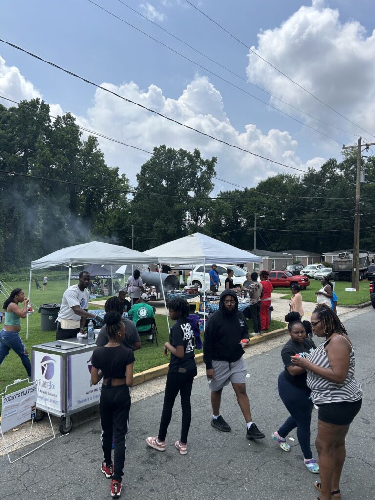 People gather at an outdoor community event with food stalls, white tents, and a cotton candy machine. Trees and parked cars line the background.