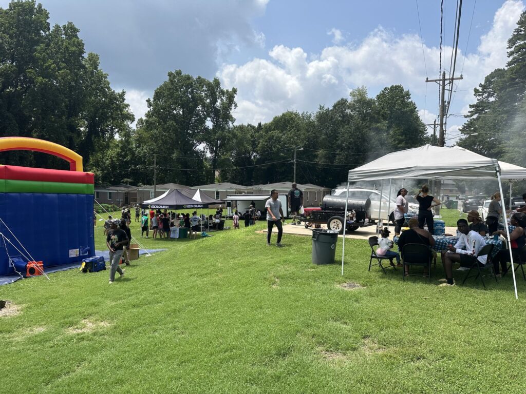 Outdoor gathering with people seated under tents and a bounce house nearby on a grassy area. Trees and clouds are visible in the background.