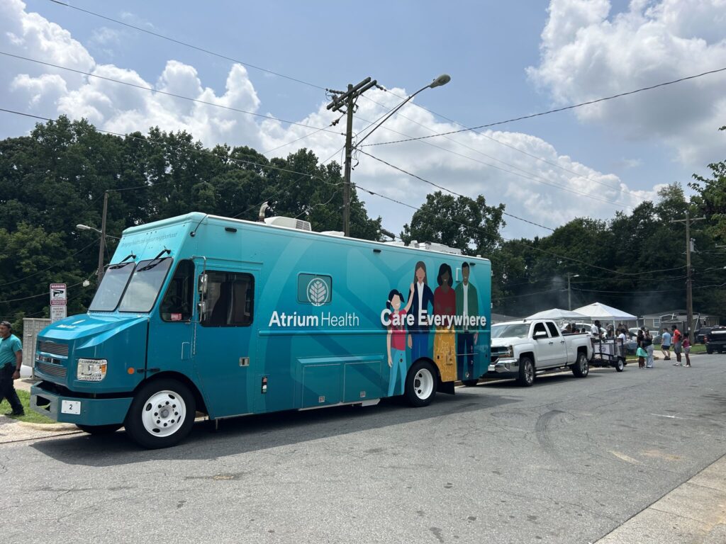 Blue Atrium Health mobile clinic truck with "Care Everywhere" slogan parked on a street near people and trees under a cloudy sky.