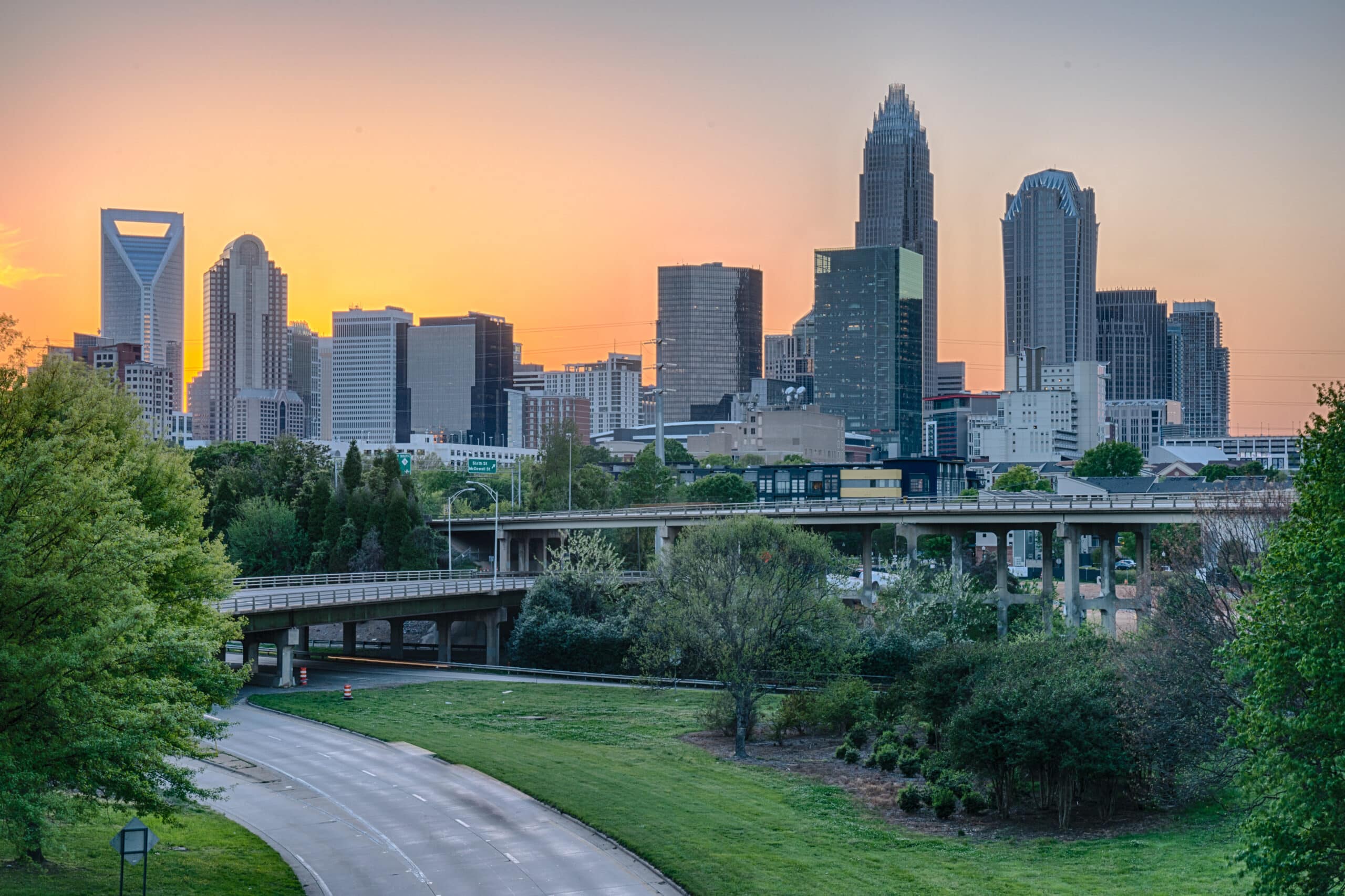 City skyline at dusk with a clear sky, featuring a mix of modern skyscrapers and a highway in the foreground, surrounded by green trees and grass.