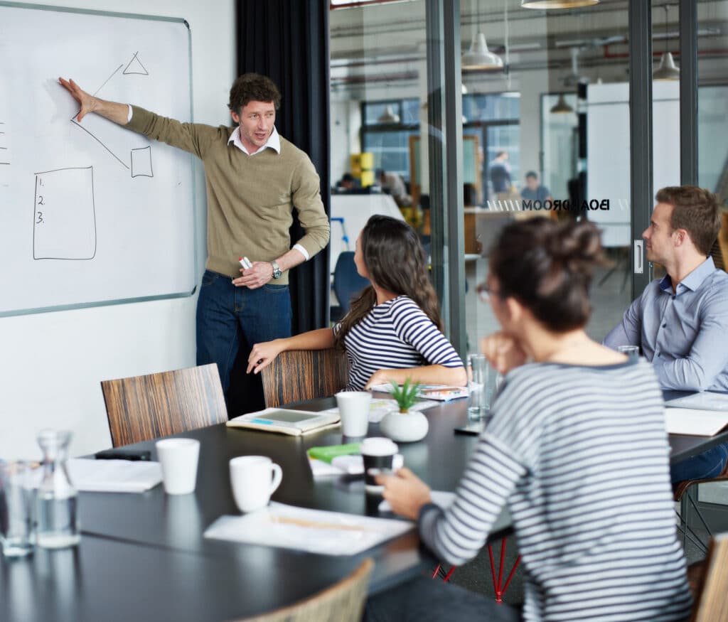 A man presents at a whiteboard in a conference room, pointing at diagrams. Three people sit at a table, listening. Coffee cups and documents are on the table.
