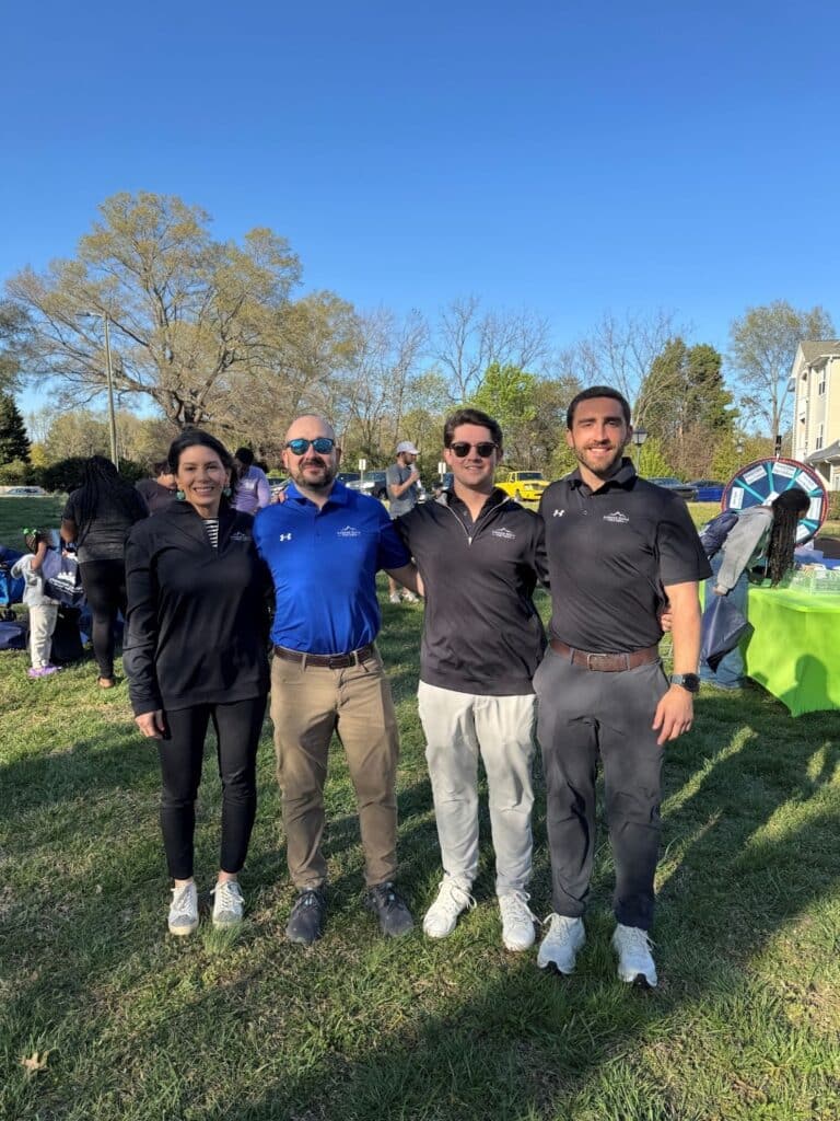 Four people stand side by side outdoors on grass, posing for a group photo. They are wearing casual athletic clothing. Trees and event tables are visible in the background.